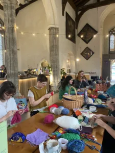 Tables inside a church, full of people knitting and crocheting. there is a bar in the background