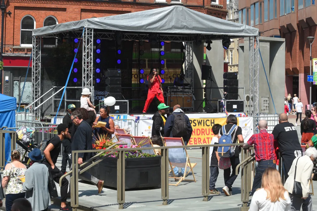 A performer in red on a covered stage in a paved square, with people gathered by an Ipswich Windrush banner
