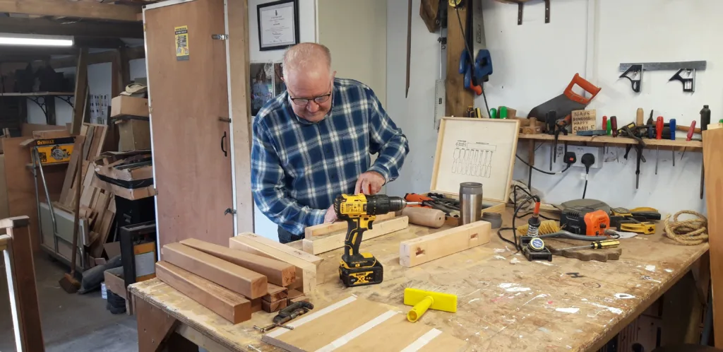 A man in a checked shirt using a cordless drill at a cluttered workbench in a wood workshop