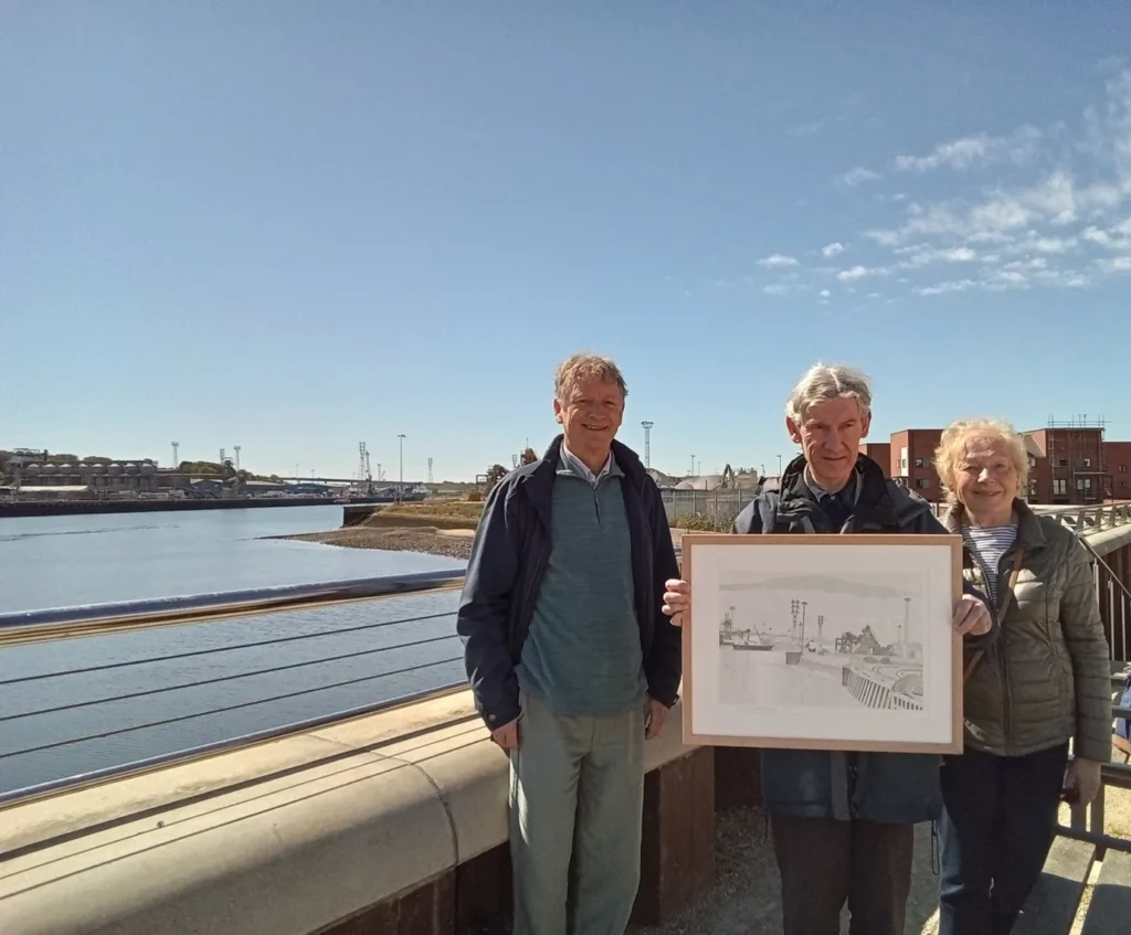 Three people standing on a sunny quayside holding a framed line drawing of a dockside scene